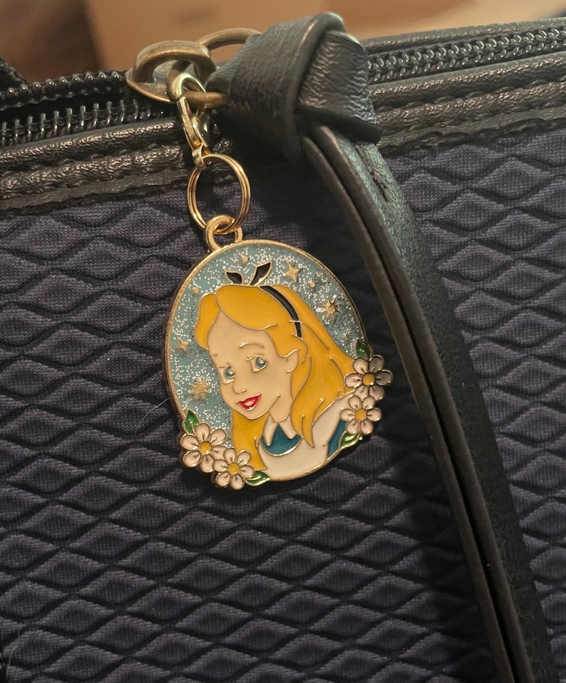 May include: A gold-coloured metal charm depicting Alice from Alice in Wonderland. The oval charm has a blue background with stars and white flowers. Alice has blonde hair and wears a blue dress. The charm is attached to a black bag.