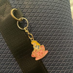 May include: A silver keyring with a charm depicting Alice in Wonderland in a pink teacup. The charm has a gold-coloured border. The background is a textured navy blue fabric.