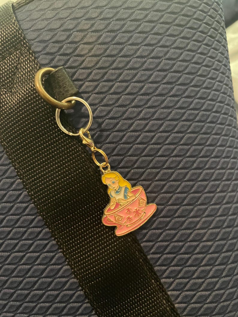 May include: A silver keyring with a charm depicting Alice in Wonderland in a pink teacup. The charm has a gold-coloured border. The background is a textured navy blue fabric.