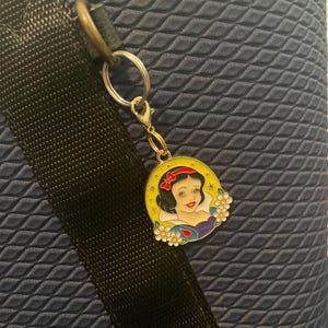 May include: A gold-colored charm featuring a portrait of Snow White with a red bow, surrounded by a yellow border and white flowers. The charm is attached to a silver keyring and clasp, set against a textured navy blue background.