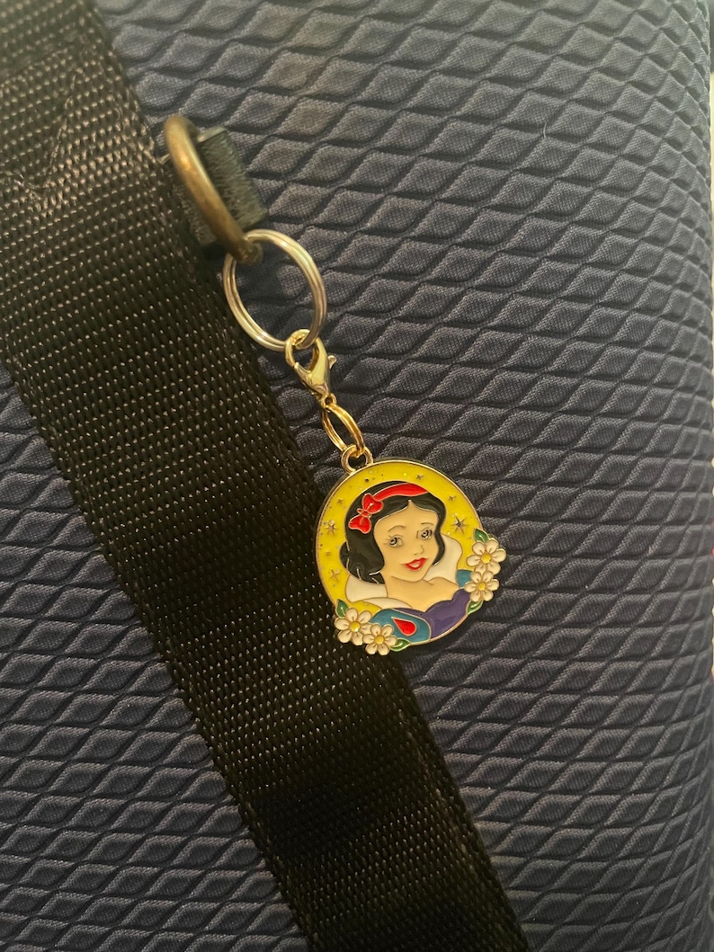 May include: A gold-colored charm featuring a portrait of Snow White with a red bow, surrounded by a yellow border and white flowers. The charm is attached to a silver keyring and clasp, set against a textured navy blue background.