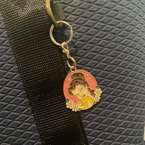 May include: A gold-colored keychain with a charm featuring a cartoon image of a woman with brown hair and a yellow dress, surrounded by white flowers, set against a pink background. The keychain is attached to a black strap and a blue textured surface.