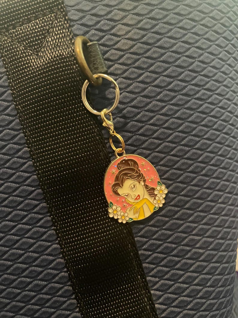 May include: A gold-colored keychain with a charm featuring a cartoon image of a woman with brown hair and a yellow dress, surrounded by white flowers, set against a pink background. The keychain is attached to a black strap and a blue textured surface.
