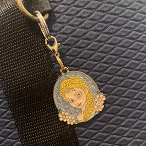 May include: A gold-colored charm featuring a cartoon image of a woman with blonde hair and a blue dress, surrounded by white flowers. The charm is attached to a silver clasp and a black strap.
