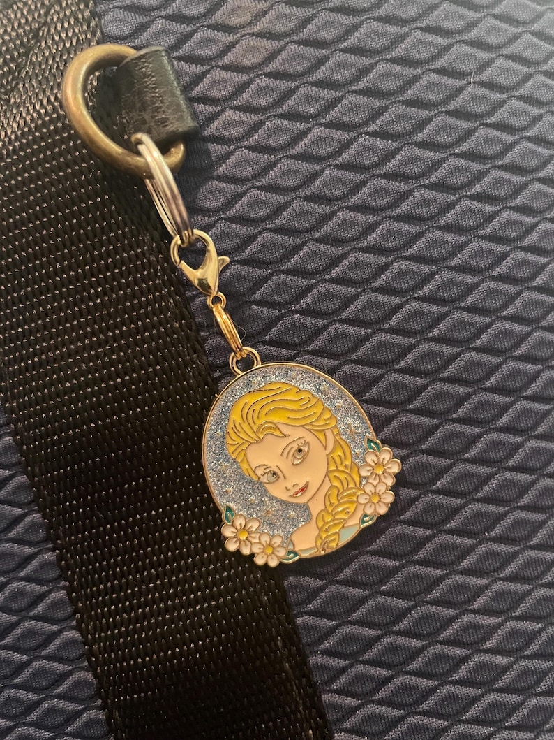 May include: A gold-colored charm featuring a cartoon image of a woman with blonde hair and a blue dress, surrounded by white flowers. The charm is attached to a silver clasp and a black strap.