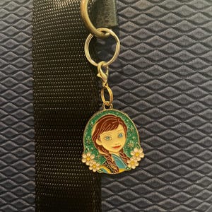 May include: A keychain featuring a cartoon image of a girl with brown braids, blue eyes, and a yellow and blue dress. The image is surrounded by white flowers with yellow centers, set against a green background. The keychain is attached to a silver keyring.