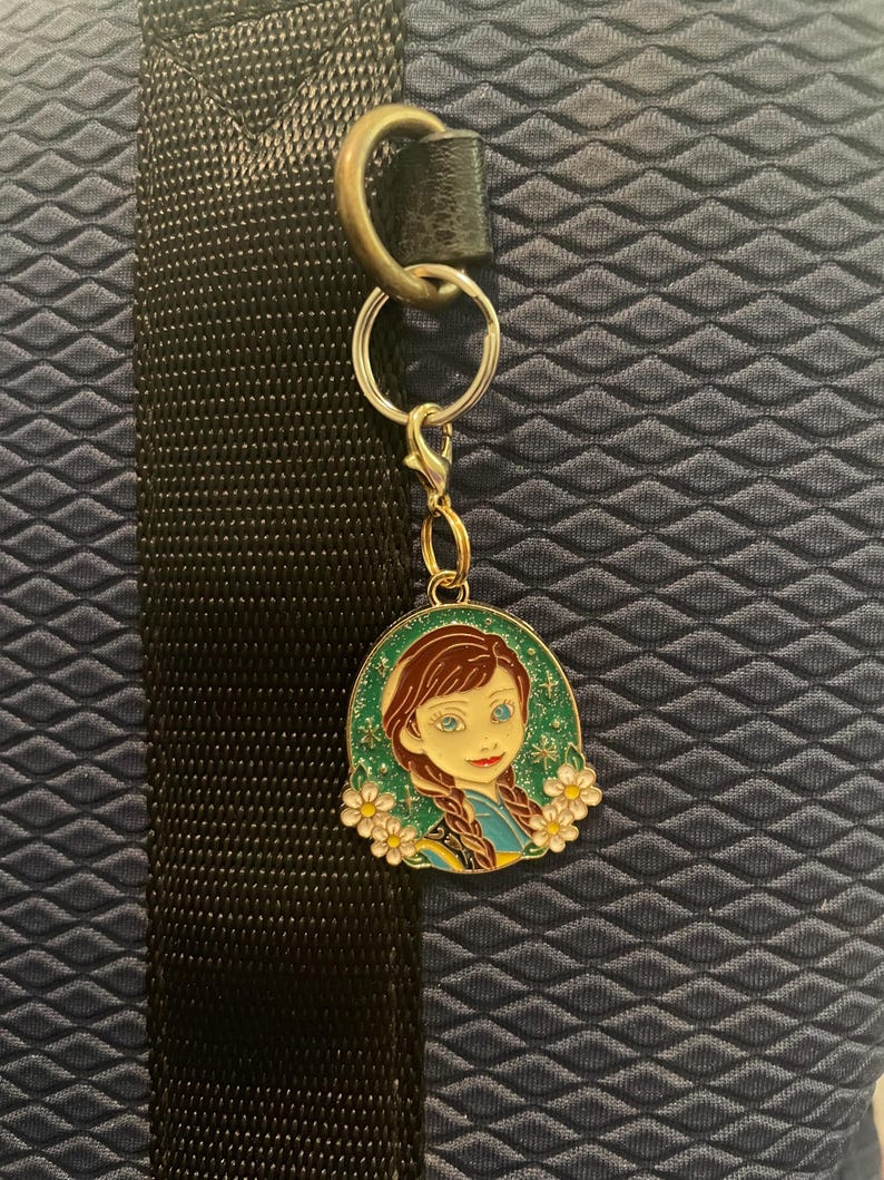 May include: A keychain featuring a cartoon image of a girl with brown braids, blue eyes, and a yellow and blue dress. The image is surrounded by white flowers with yellow centers, set against a green background. The keychain is attached to a silver keyring.