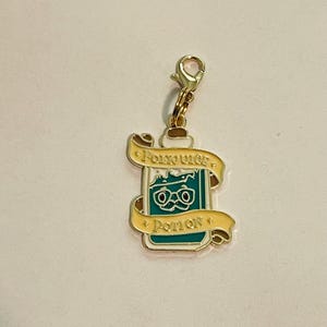 Puede incluir: Un charm de color dorado con un cierre de mosquetón. El charm presenta un diseño de botella de poción turquesa con una pancarta que dice "Potion". La botella de poción tiene un par de gafas. La pancarta es amarilla con un contorno dorado.