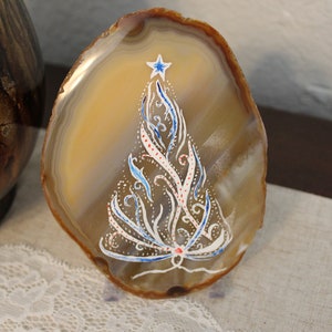 Puede incluir: Una rebanada de ágata decorativa con un diseño de árbol de Navidad pintado a mano. El árbol es blanco con detalles azules y rojos, coronado por una estrella azul. El ágata tiene tonos naturales marrones y beige.