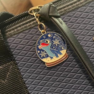 May include: A gold-colored charm featuring a blue lizard inside a snow globe design with snowflakes. The charm is attached to a navy blue bag with a zipper. The lizard has a red tongue and is set against a blue background.