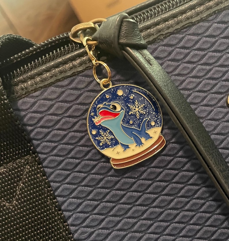 May include: A gold-colored charm featuring a blue lizard inside a snow globe design with snowflakes. The charm is attached to a navy blue bag with a zipper. The lizard has a red tongue and is set against a blue background.