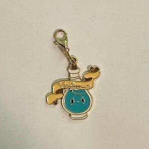 Puede incluir: Un dije dorado con un diseño de botella de poción azul. La botella tiene una cara linda y está rodeada por una pancarta con el texto "Veritaserum". El dije está sujeto a un cierre dorado.