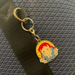 May include: A circular gold-coloured keychain charm depicting a cartoon girl with red hair, a blue top, and white flowers. The charm is attached to a gold-coloured clasp and a silver keyring. The background is a dark blue, textured surface.