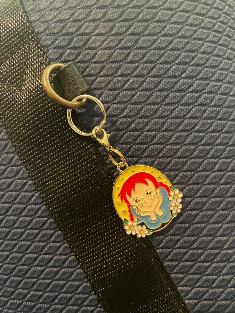 May include: A circular gold-coloured keychain charm depicting a cartoon girl with red hair, a blue top, and white flowers. The charm is attached to a gold-coloured clasp and a silver keyring. The background is a dark blue, textured surface.