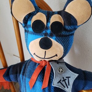 Puede incluir: Mu&ntilde;eca de oso hecha a mano con un patr&oacute;n de cuadros azules y negros. El oso tiene orejas color canela, ojos negros y una sonrisa cosida. Lleva una bufanda roja y una insignia gris con una estrella y las letras "TTT".