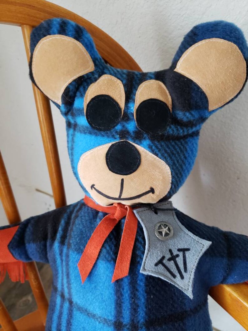 Puede incluir: Mu&ntilde;eca de oso hecha a mano con un patr&oacute;n de cuadros azules y negros. El oso tiene orejas color canela, ojos negros y una sonrisa cosida. Lleva una bufanda roja y una insignia gris con una estrella y las letras "TTT".