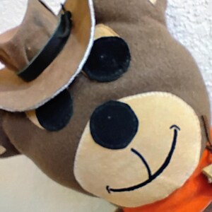 Puede incluir: Un oso de peluche marr&oacute;n con un sombrero de vaquero, ojos negros y una cara sonriente. El oso tiene un hocico beige y un pa&ntilde;uelo naranja. El sombrero de vaquero es marr&oacute;n con una banda negra.