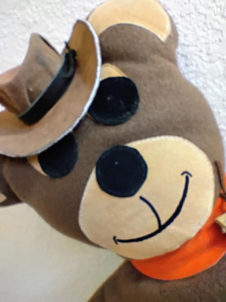 Puede incluir: Un oso de peluche marr&oacute;n con un sombrero de vaquero, ojos negros y una cara sonriente. El oso tiene un hocico beige y un pa&ntilde;uelo naranja. El sombrero de vaquero es marr&oacute;n con una banda negra.