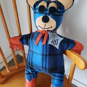 Puede incluir: Un oso de peluche hecho a mano con un patr&oacute;n de cuadros azules y negros. El oso tiene orejas color beige, ojos negros y detalles naranjas en los brazos y las botas. Lleva una cinta roja y una insignia con las letras "T T".