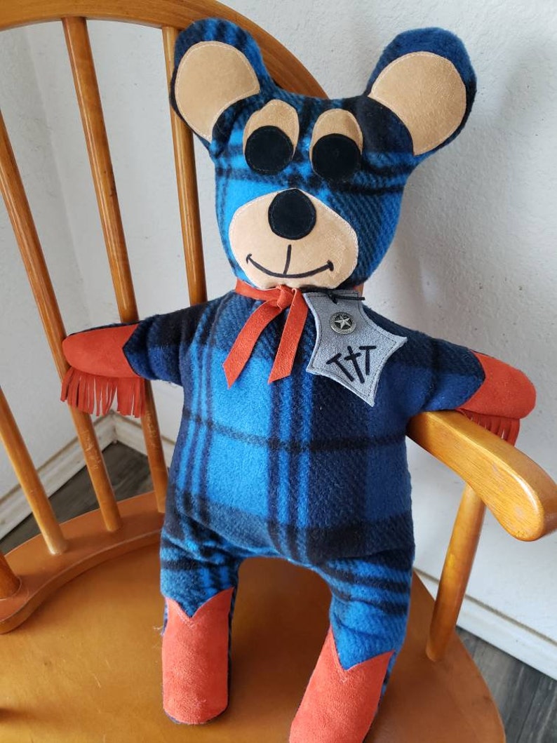 Puede incluir: Un oso de peluche hecho a mano con un patr&oacute;n de cuadros azules y negros. El oso tiene orejas color beige, ojos negros y detalles naranjas en los brazos y las botas. Lleva una cinta roja y una insignia con las letras "T T".