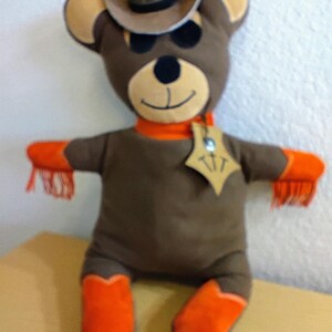 Puede incluir: Un oso de peluche marr&oacute;n con sombrero de vaquero, bufanda naranja y botas naranjas. El oso tiene una placa de sheriff y flecos naranjas en los brazos. La cara del oso tiene una expresi&oacute;n sonriente.