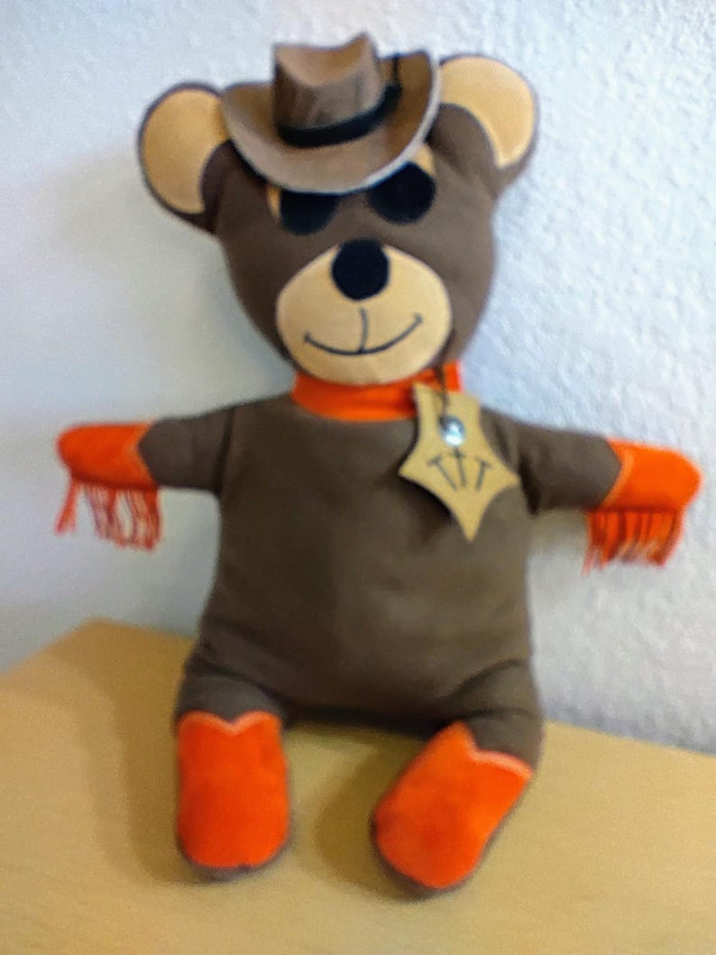 Puede incluir: Un oso de peluche marr&oacute;n con sombrero de vaquero, bufanda naranja y botas naranjas. El oso tiene una placa de sheriff y flecos naranjas en los brazos. La cara del oso tiene una expresi&oacute;n sonriente.