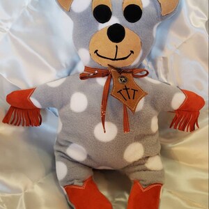 Puede incluir: Un oso de peluche hecho a mano con tela gris y lunares blancos. El oso tiene orejas marrones, hocico marr&oacute;n y ojos negros. Lleva botas de vaquero naranjas y tiene flecos naranjas en los brazos. Una etiqueta de cuero marr&oacute;n con texto cuelga de su cuello.