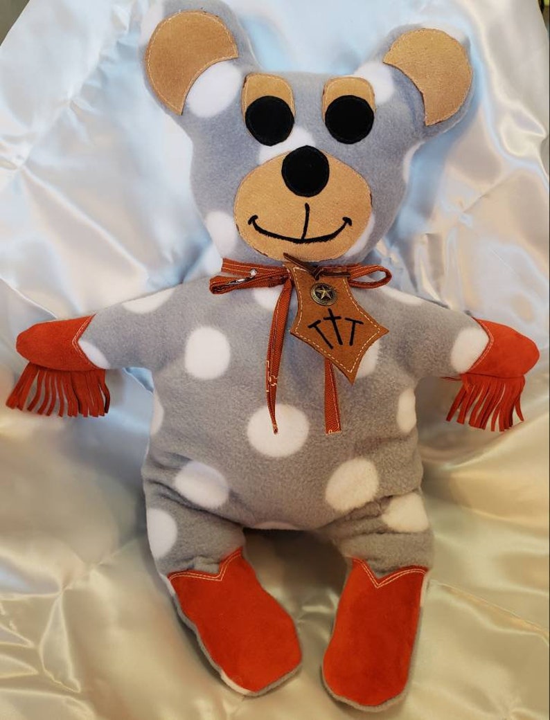 Puede incluir: Un oso de peluche hecho a mano con tela gris y lunares blancos. El oso tiene orejas marrones, hocico marr&oacute;n y ojos negros. Lleva botas de vaquero naranjas y tiene flecos naranjas en los brazos. Una etiqueta de cuero marr&oacute;n con texto cuelga de su cuello.