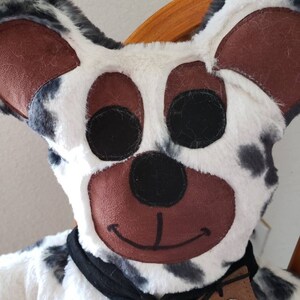 Puede incluir: Un juguete de peluche con forma de perro con un patr&oacute;n de manchas blancas y negras. El perro tiene grandes orejas marrones, ojos negros y una nariz marr&oacute;n. Lleva una bufanda negra y una etiqueta de cuero marr&oacute;n con un emblema de estrella.