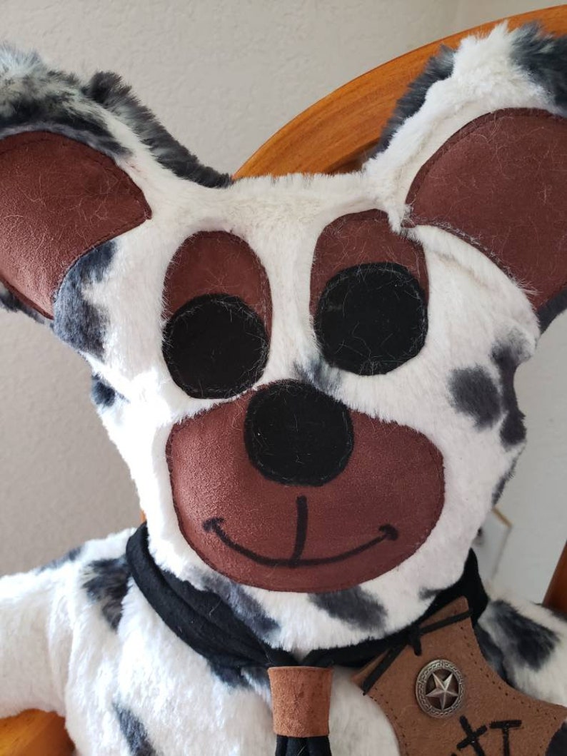 Puede incluir: Un juguete de peluche con forma de perro con un patr&oacute;n de manchas blancas y negras. El perro tiene grandes orejas marrones, ojos negros y una nariz marr&oacute;n. Lleva una bufanda negra y una etiqueta de cuero marr&oacute;n con un emblema de estrella.