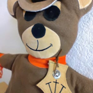 Puede incluir: Mu&ntilde;eca de oso de peluche marr&oacute;n con sombrero de vaquero con banda negra y pa&ntilde;uelo naranja. El oso tiene ojos negros y hocico beige. Una etiqueta con las letras "TTT" cuelga de su cuello.