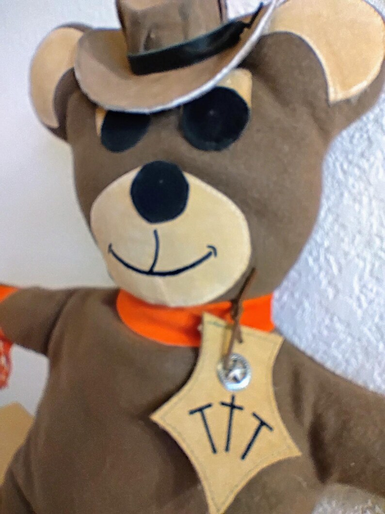 Puede incluir: Mu&ntilde;eca de oso de peluche marr&oacute;n con sombrero de vaquero con banda negra y pa&ntilde;uelo naranja. El oso tiene ojos negros y hocico beige. Una etiqueta con las letras "TTT" cuelga de su cuello.