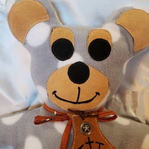 Puede incluir: Un oso de peluche hecho a mano con cuerpo gris y lunares blancos. El oso tiene orejas marrones, hocico marr&oacute;n y ojos negros. Lleva una etiqueta de cuero marr&oacute;n con una estrella y las letras "TTT". Una cinta roja est&aacute; atada alrededor de su cuello.