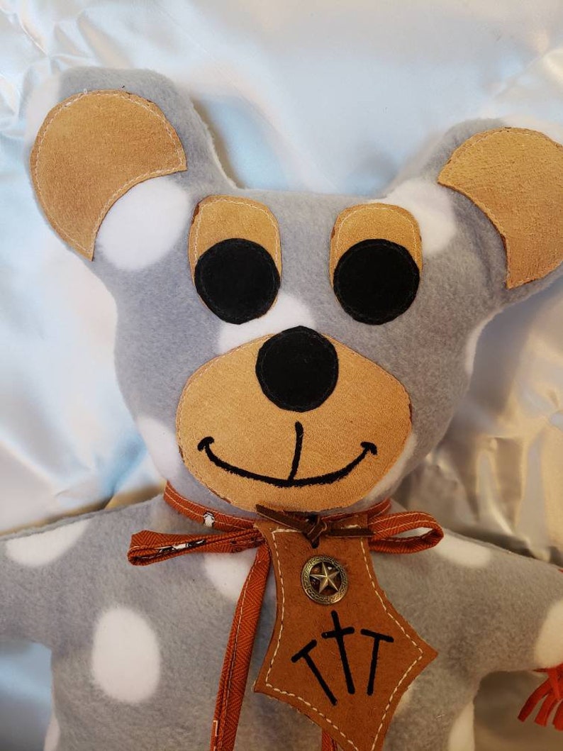 Puede incluir: Un oso de peluche hecho a mano con cuerpo gris y lunares blancos. El oso tiene orejas marrones, hocico marr&oacute;n y ojos negros. Lleva una etiqueta de cuero marr&oacute;n con una estrella y las letras "TTT". Una cinta roja est&aacute; atada alrededor de su cuello.