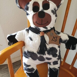 Puede incluir: Un oso de peluche hecho a mano con un patr&oacute;n de vaca de manchas negras y grises sobre piel blanca. El oso tiene cara, orejas y ojos negros marrones. Lleva una bufanda negra y una etiqueta de cuero con las iniciales "XT".