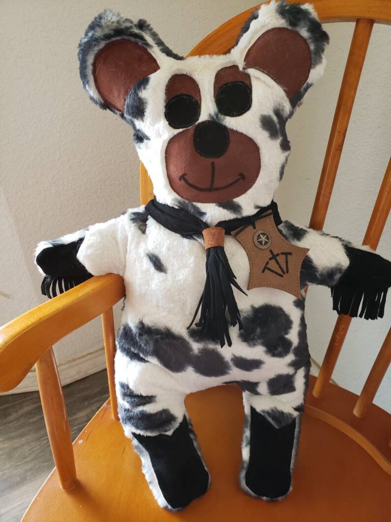 Puede incluir: Un oso de peluche hecho a mano con un patr&oacute;n de vaca de manchas negras y grises sobre piel blanca. El oso tiene cara, orejas y ojos negros marrones. Lleva una bufanda negra y una etiqueta de cuero con las iniciales "XT".
