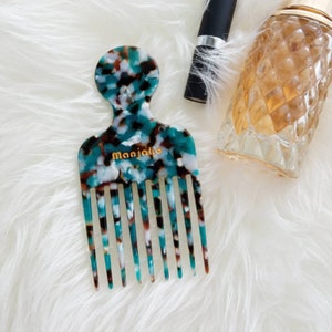 Peut inclure: Un peigne à motifs turquoise, brun et blanc avec une large poignée et le mot "Manjaka" imprimé en lettres dorées.