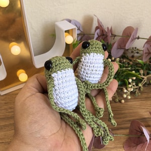 May include: Two handmade crochet frog toys. Each frog has a green head and legs, a white body, and black eyes. The frogs are held in a hand, with a decorative background featuring a gold-colored light-up letter and faux foliage.
