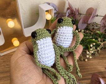 May include: Two handmade crochet frog toys. Each frog has a green head and legs, a white body, and black eyes. The frogs are held in a hand, with a decorative background featuring a gold-colored light-up letter and faux foliage.