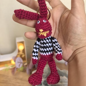 May include: A handmade crochet keychain featuring a red bunny with long ears, white eyes, and a yellow mouth. The bunny wears a black and white checkered shirt and red pants. The keychain is attached to a gold-colored ring.