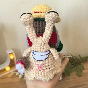 Puede incluir: Un juguete de caracol de ganchillo hecho a mano en beige, con una cara sonriente y una cruz rosa en el cuerpo. El caracol tiene un sombrero amarillo y una concha colorida. El juguete se sostiene en una mano.