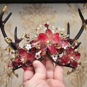 May include: A decorative headpiece featuring black antler-shaped elements adorned with red floral accents, pearls, and gold-colored embellishments. The headpiece has dangling chains and is held by a hand.