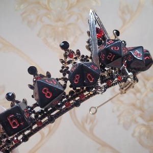 Puede incluir: Una corona negra con acentos de dados rojos. La corona est&aacute; hecha de metal y tiene un alambre plateado unido a ella.