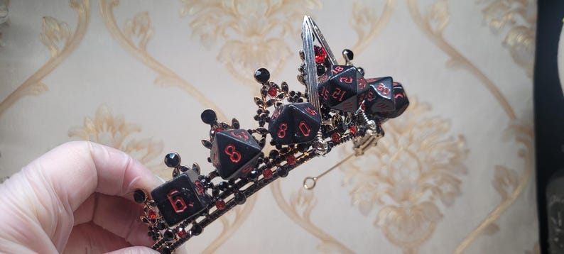 Puede incluir: Una corona negra con acentos de dados rojos. La corona est&aacute; hecha de metal y tiene un alambre plateado unido a ella.