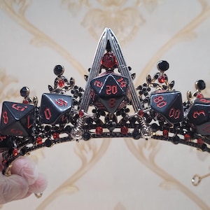 Puede incluir: Una corona negra y roja con una base dorada y un dise&ntilde;o de dados. La corona est&aacute; decorada con gemas rojas y detalles en metal negro.