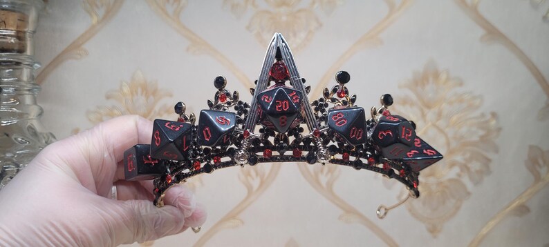 Puede incluir: Una corona negra y roja con una base dorada y un dise&ntilde;o de dados. La corona est&aacute; decorada con gemas rojas y detalles en metal negro.