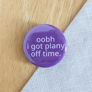 May include: A circular, purple button featuring the text "oobh i got plany off time" in white. The button is set against a wooden surface and a grey fabric. This is a pin badge.