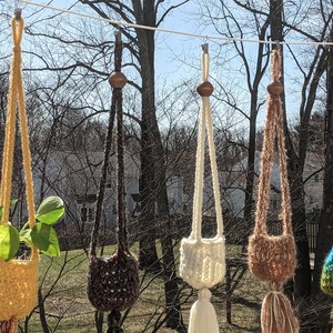 May include: Six macrame plant hangers with wooden beads hanging from a clothesline. The hangers are in various colors, including blue, yellow, brown, white, and brown. One of the hangers has a green plant inside.