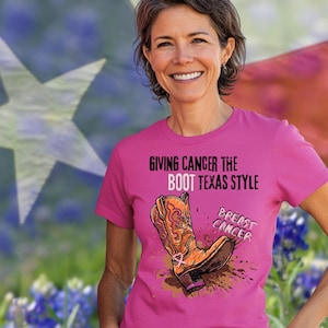 Könnte beinhalten: Pinkes T-Shirt mit einem Grafikdruck einer Cowboystiefel, die ein rosa Band mit dem Text "Giving Cancer the Boot Texas Style" und "Breast Cancer" unter dem Stiefel treten.