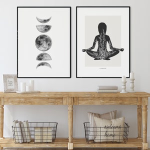 Puede incluir: Dos impresiones enmarcadas en negro en una pared blanca sobre una mesa consola de madera. La impresi&oacute;n de la izquierda muestra una ilustraci&oacute;n en blanco y negro de la luna en diferentes fases. La impresi&oacute;n de la derecha muestra una silueta negra de una persona en una postura de yoga con un dise&ntilde;o floral dentro de la silueta. El texto "SAMASITI" est&aacute; debajo de la imagen. La mesa consola tiene dos cestas de alambre negro en el estante inferior. La cesta de la izquierda contiene libros y la cesta de la derecha contiene una almohada con el texto "welcome home".